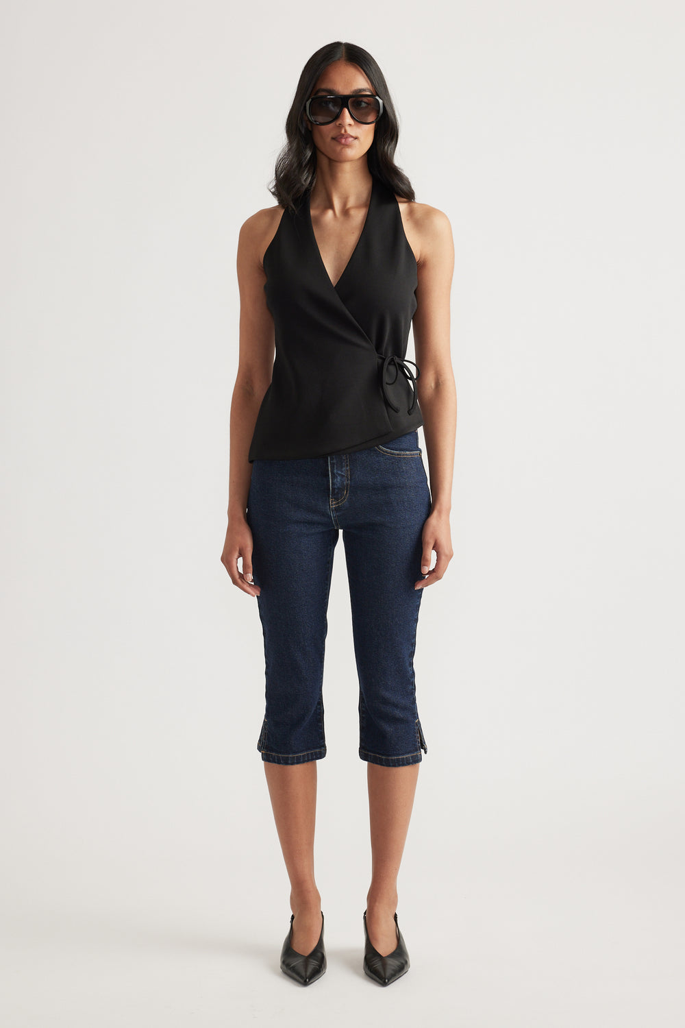 Woman wearing a black sleeveless wrap top with side tie, dark cropped jeans, sunglasses and pointed heels