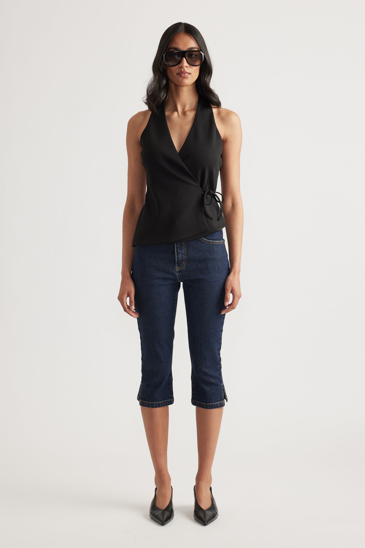 Woman wearing a black sleeveless wrap top with side tie, dark cropped jeans, sunglasses and pointed heels