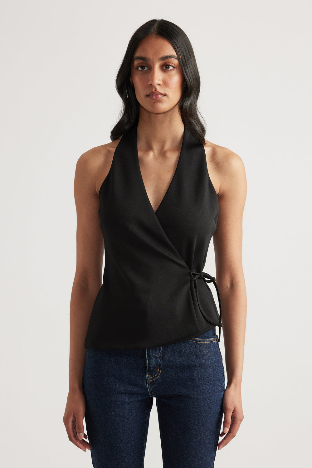 Woman wearing a black wrap halter neck top with side waist tie and V neck, styled with dark blue jeans