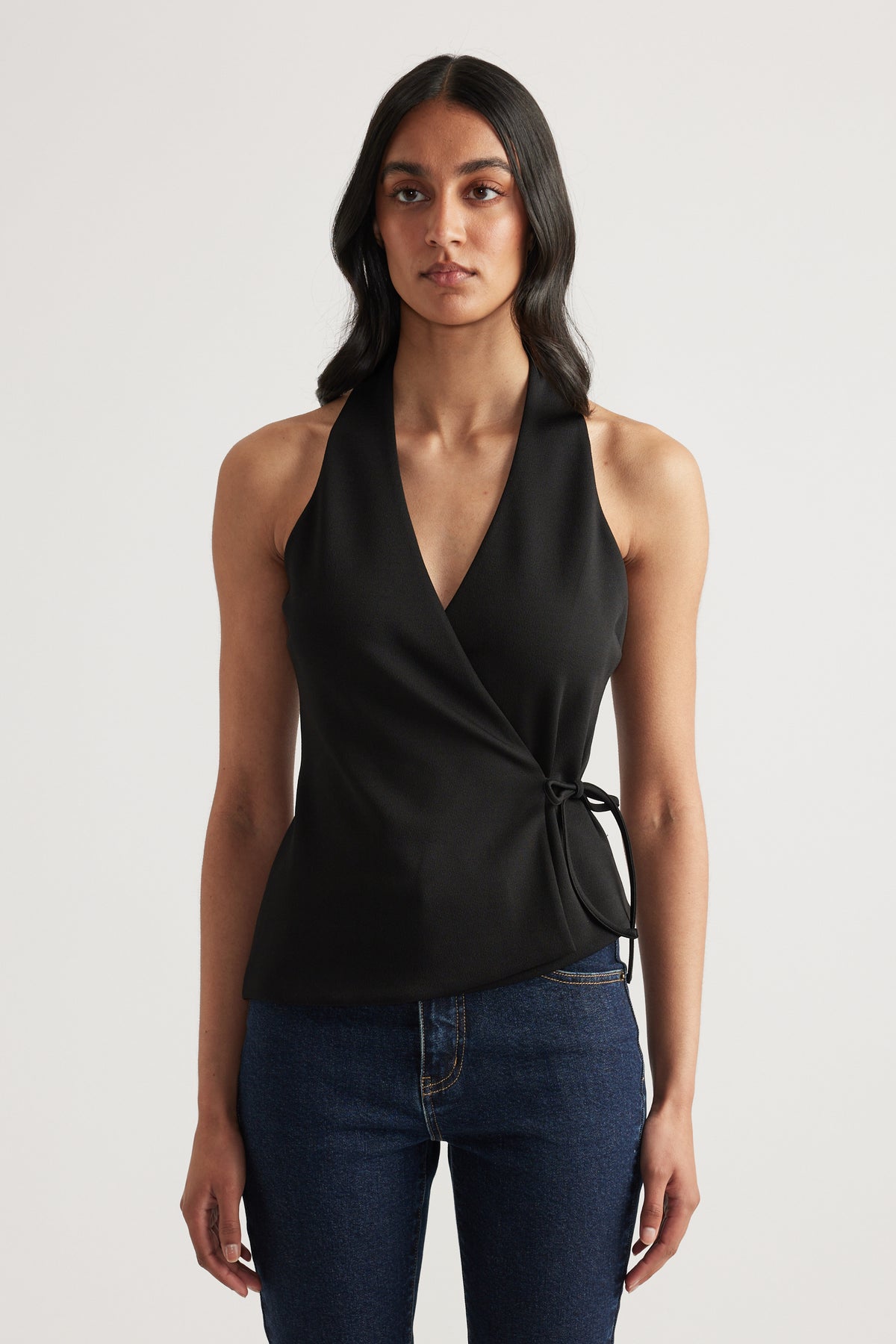 Woman wearing a black wrap halter neck top with side waist tie and V neck, styled with dark blue jeans