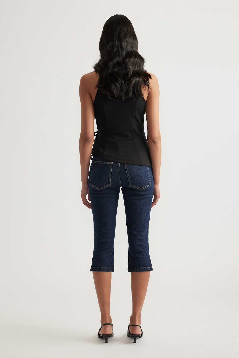 Back view of woman wearing a black sleeveless wrap halter top with dark cropped jeans and black heels