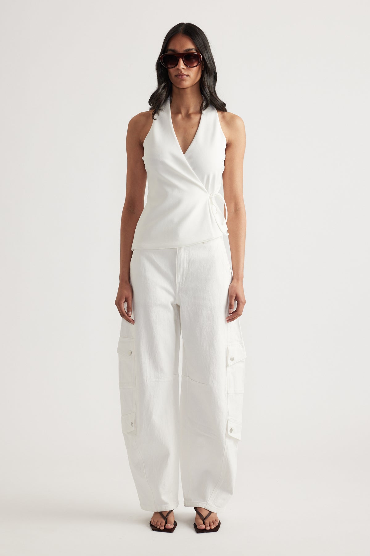 Woman wearing a white sleeveless wrap halter top with side tie, white cargo pants, sunglasses and sandals