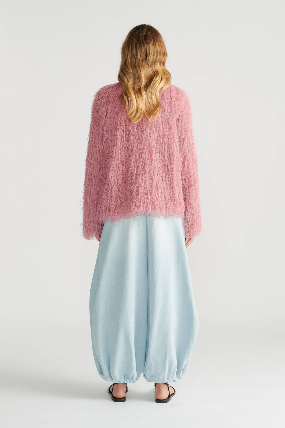 Woman wearing a soft pink fringed jacket paired with light blue balloon pants and black sandals, back view.