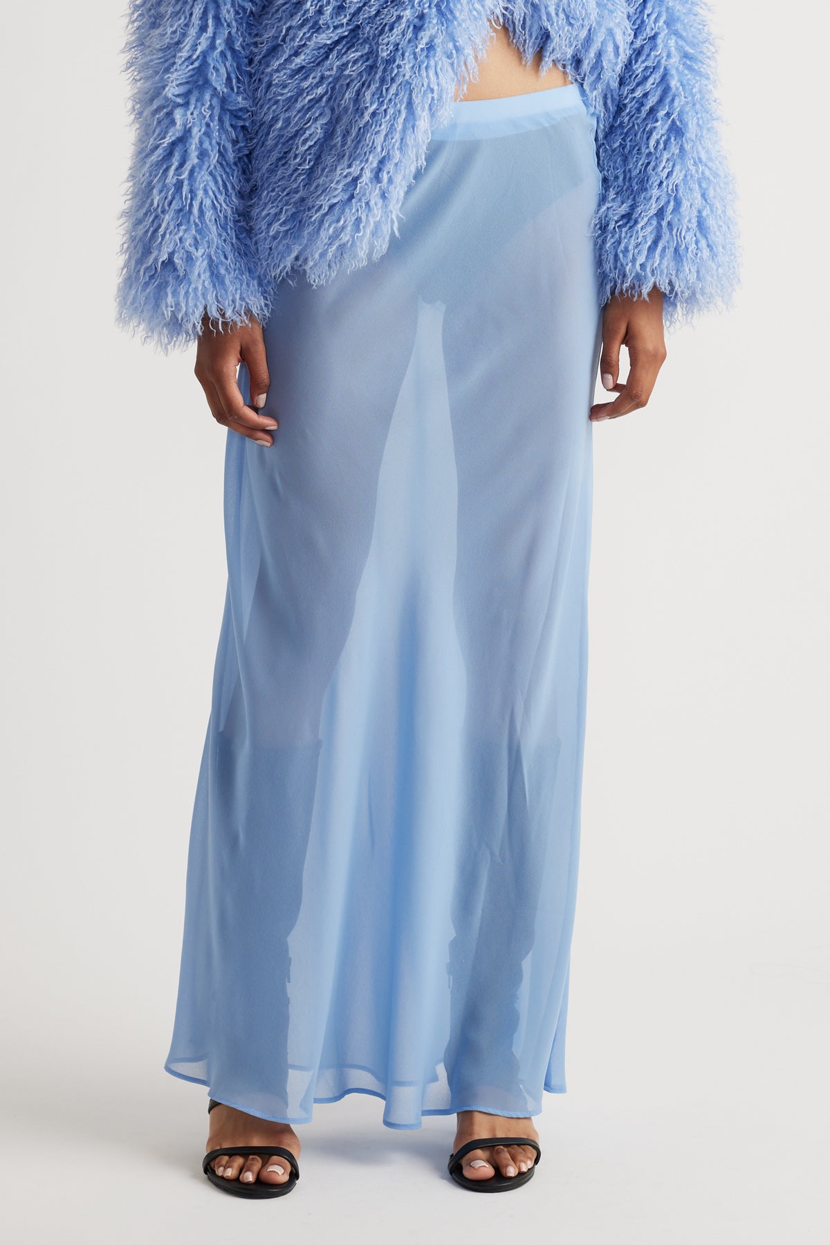 Sheer light blue maxi skirt with a flowy silhouette paired with a textured, fluffy cropped jacket and black sandals.