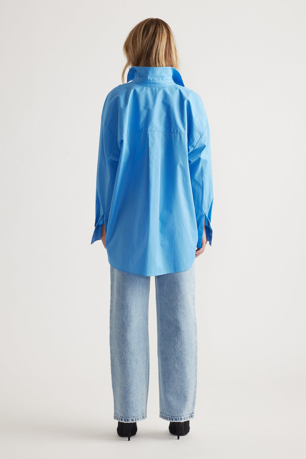 Woman wearing a relaxed fit oversized blue shirt paired with straight-leg light wash denim jeans and black heels, back view.