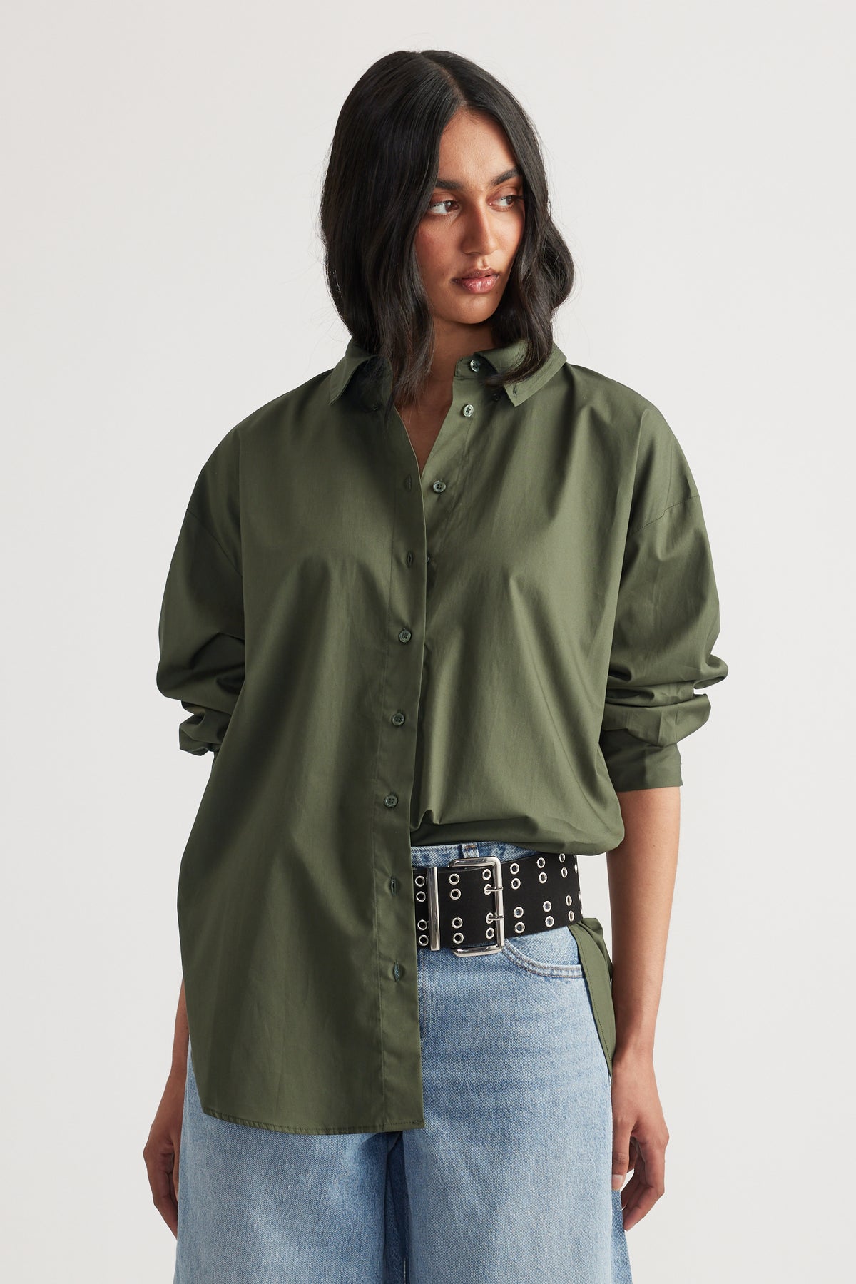 Woman wearing an oversized olive green button-up shirt styled with light blue wide-leg jeans and a black grommet belt