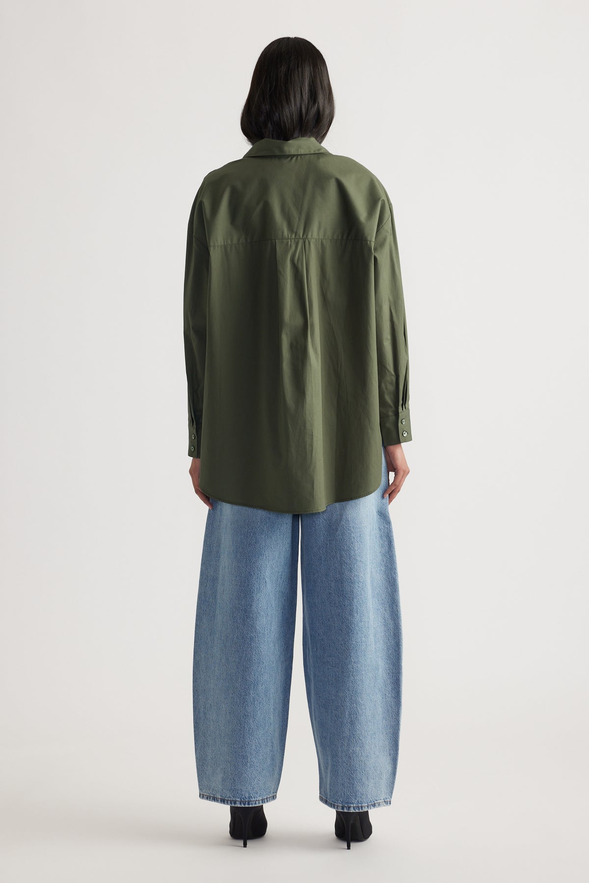 Woman seen from back wearing oversized olive green shirt with button cuffs and wide leg light blue denim jeans