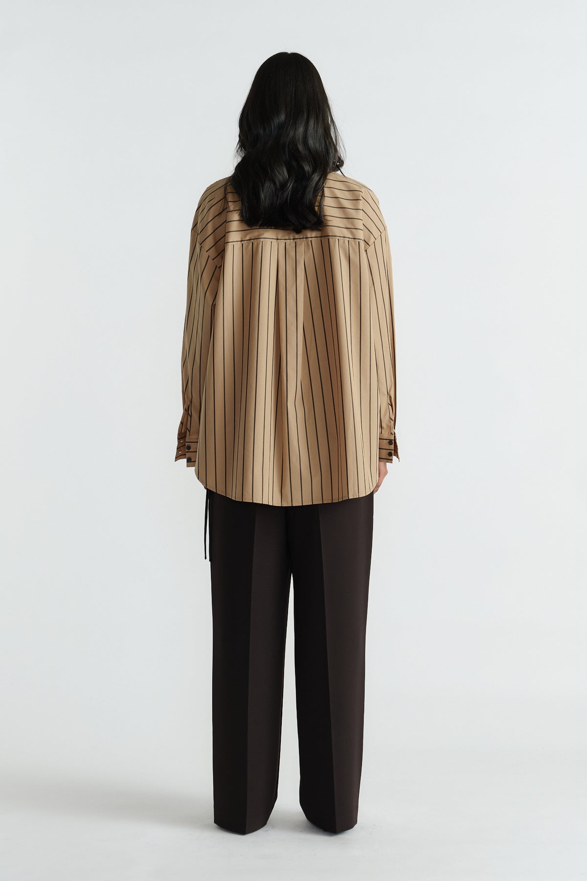 Woman shown from behind wearing a loose tan striped shirt with black wide-leg trousers against white background.