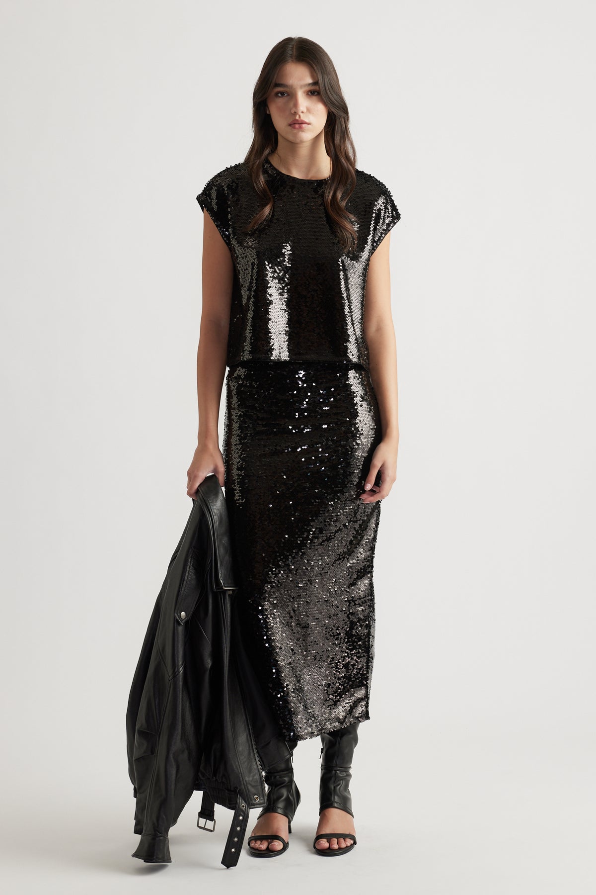 Woman wearing a black sequin shell top paired with a matching sequin skirt, holding a leather jacket and black heels.