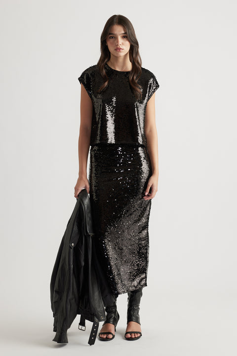 Woman wearing a black sequin shell top paired with a matching sequin skirt, holding a leather jacket and black heels.