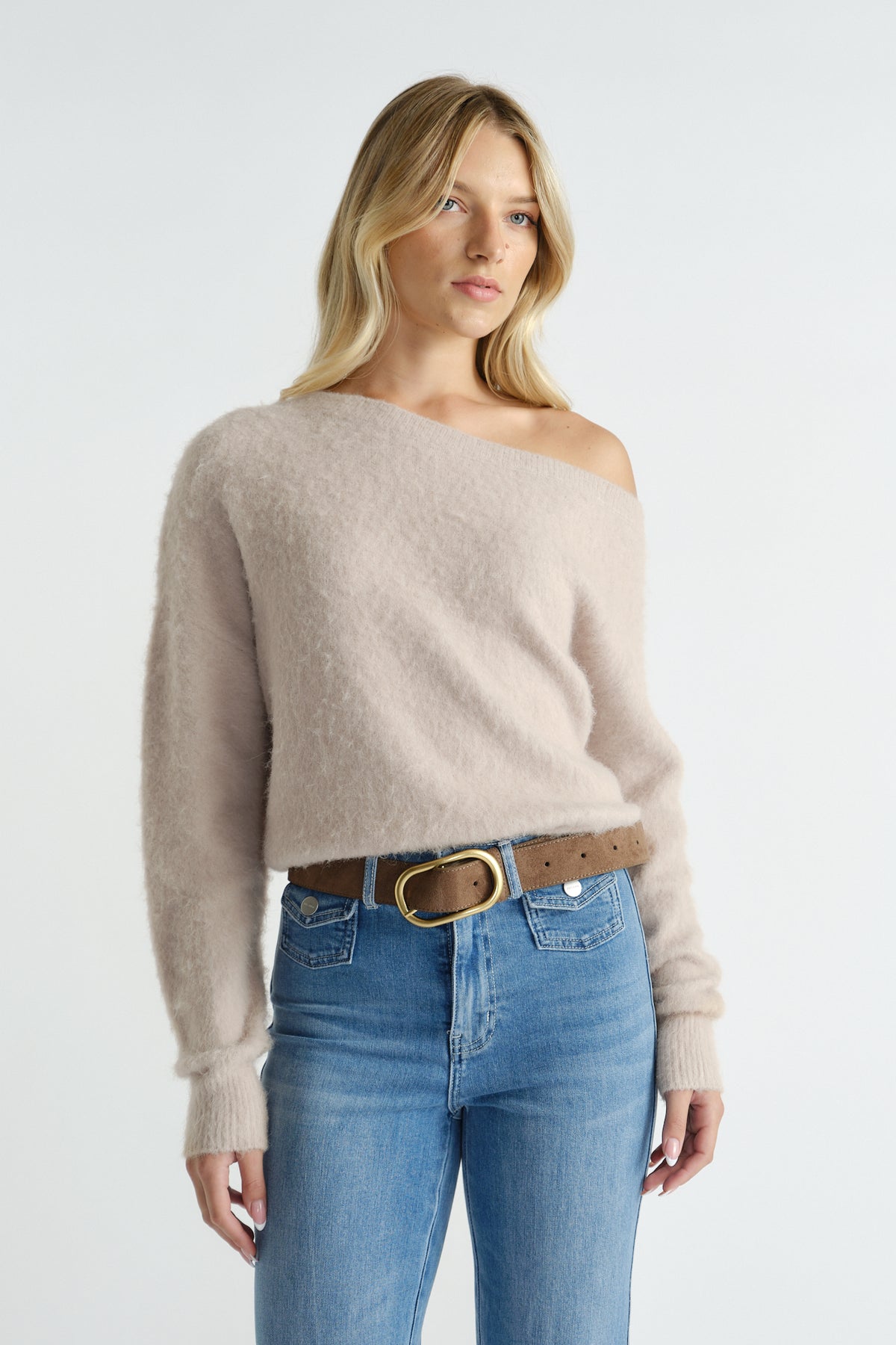 Woman wearing a soft beige off-shoulder knit paired with high-waisted blue jeans and a brown belt with gold buckle
