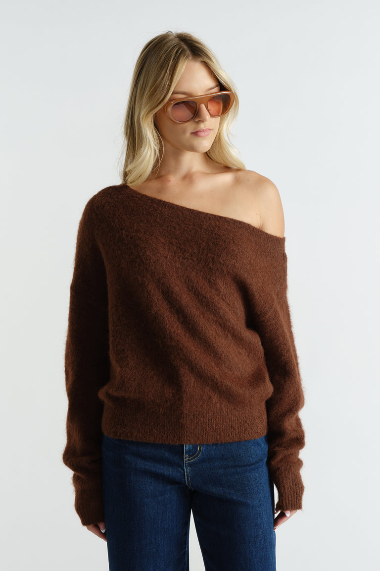 Woman wearing a soft brown off-shoulder knit sweater paired with dark blue jeans and brown-tinted sunglasses