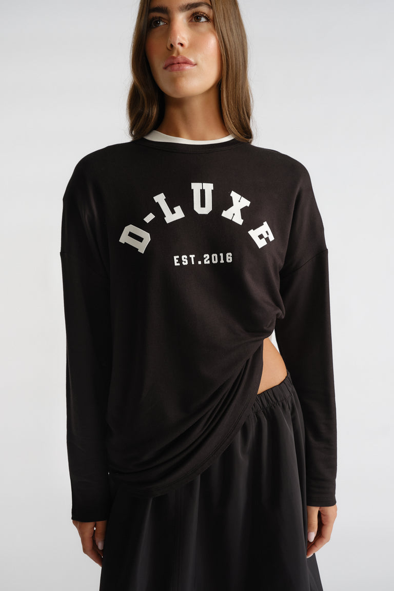 Woman wearing a black long sleeve top with white bold logo detail paired with a flowing black skirt