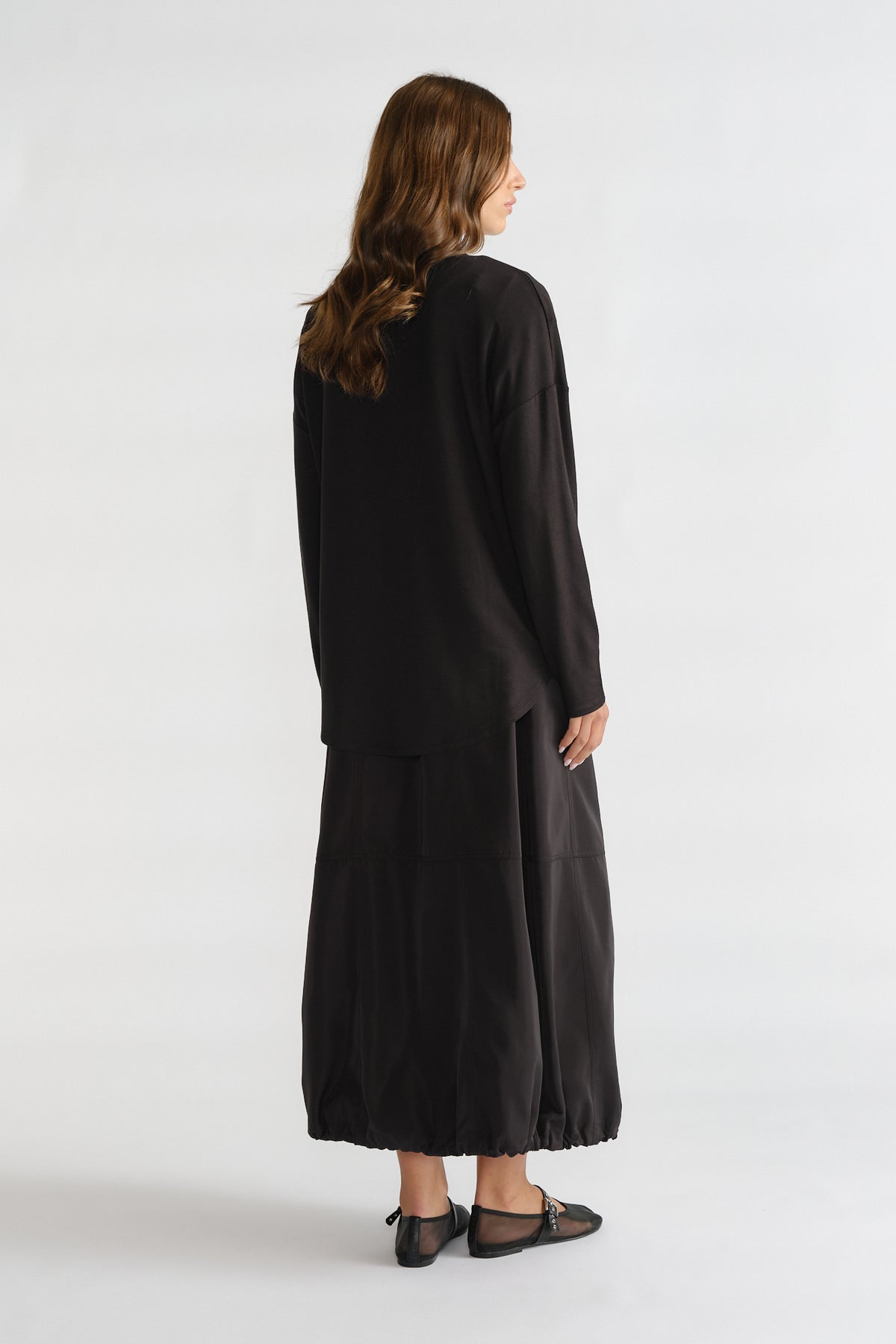Woman shown from behind wearing a relaxed loose black long sleeve top paired with a flowy black skirt and flats