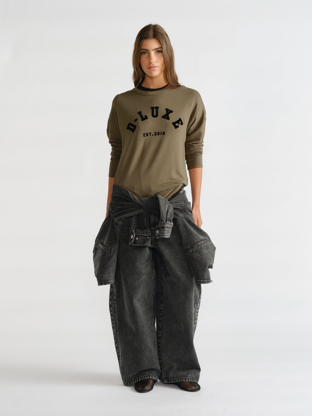 Woman wearing an olive green long sleeve top with logo detail paired with wide-leg faded black denim jeans and a tied jacket around the waist
