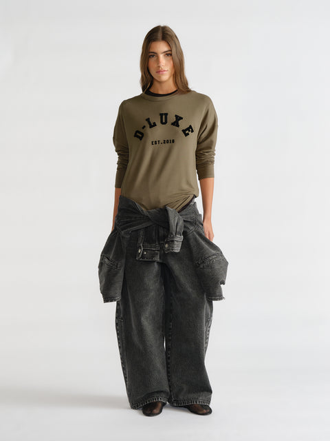 Woman wearing an olive green long sleeve top with logo detail paired with wide-leg faded black denim jeans and a tied jacket around the waist