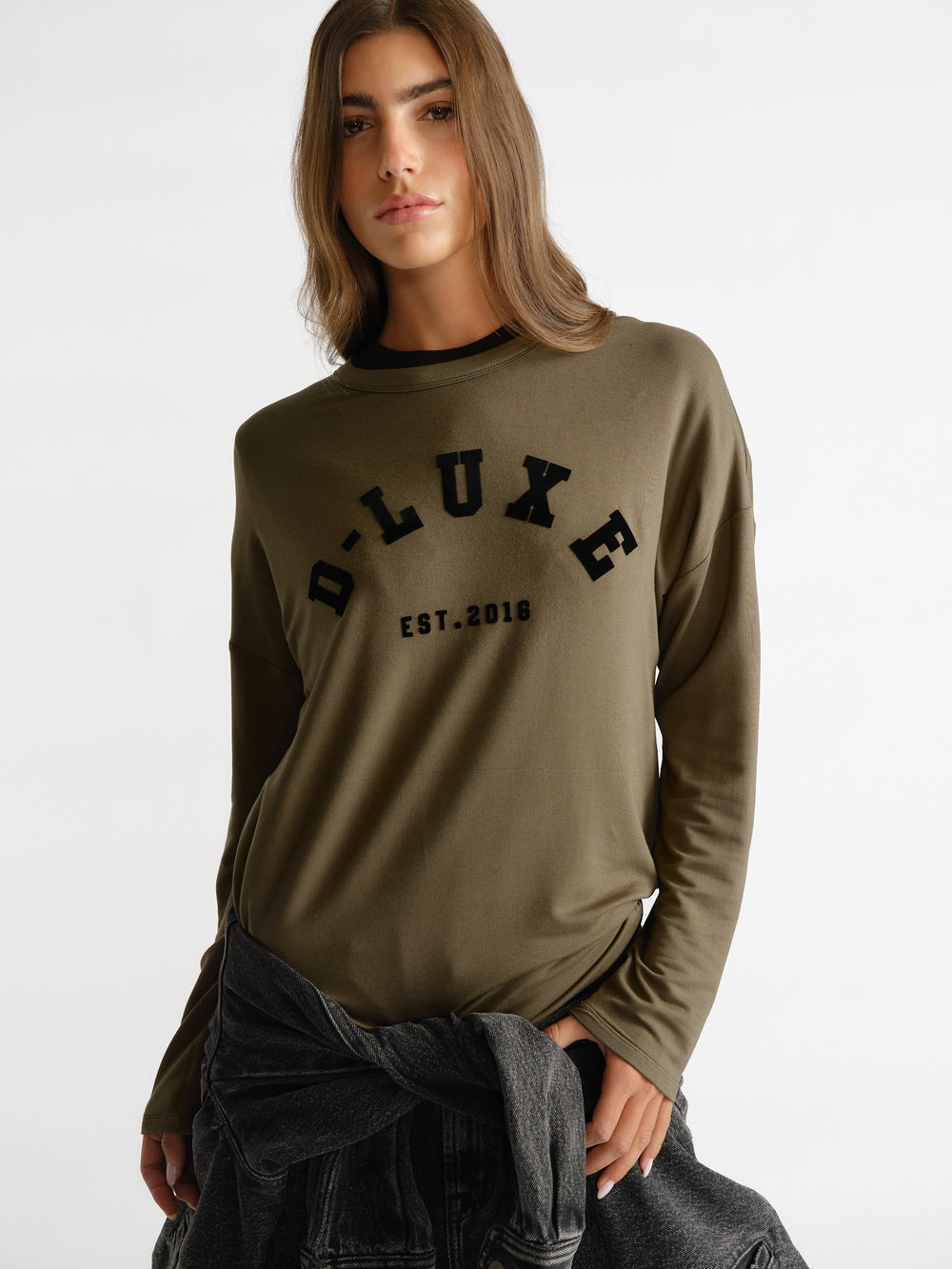 Woman wearing an olive green long sleeve top with black D-Luxe logo paired with a tied dark denim jacket at the waist