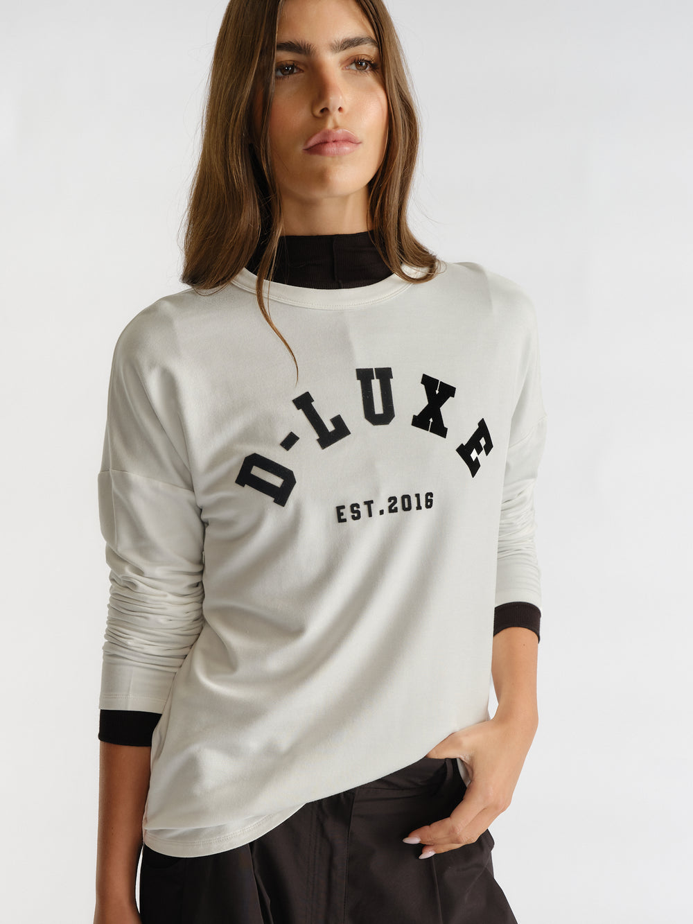 Woman wearing an ivory long sleeve top with bold black logo detail layered over a black turtleneck and paired with dark pants