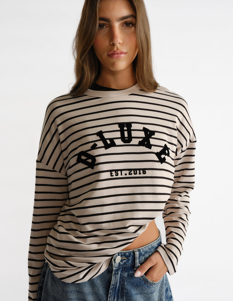 Woman wearing a beige and black striped long sleeve top with bold logo print, styled with casual blue jeans