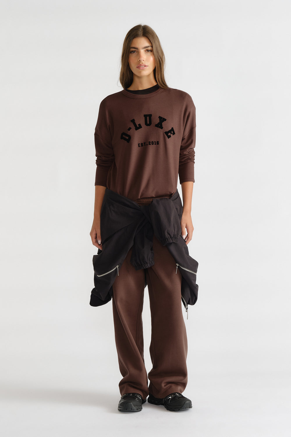 Woman wearing a relaxed brown long sleeve logo top with matching loose pants and a black jacket tied at the waist