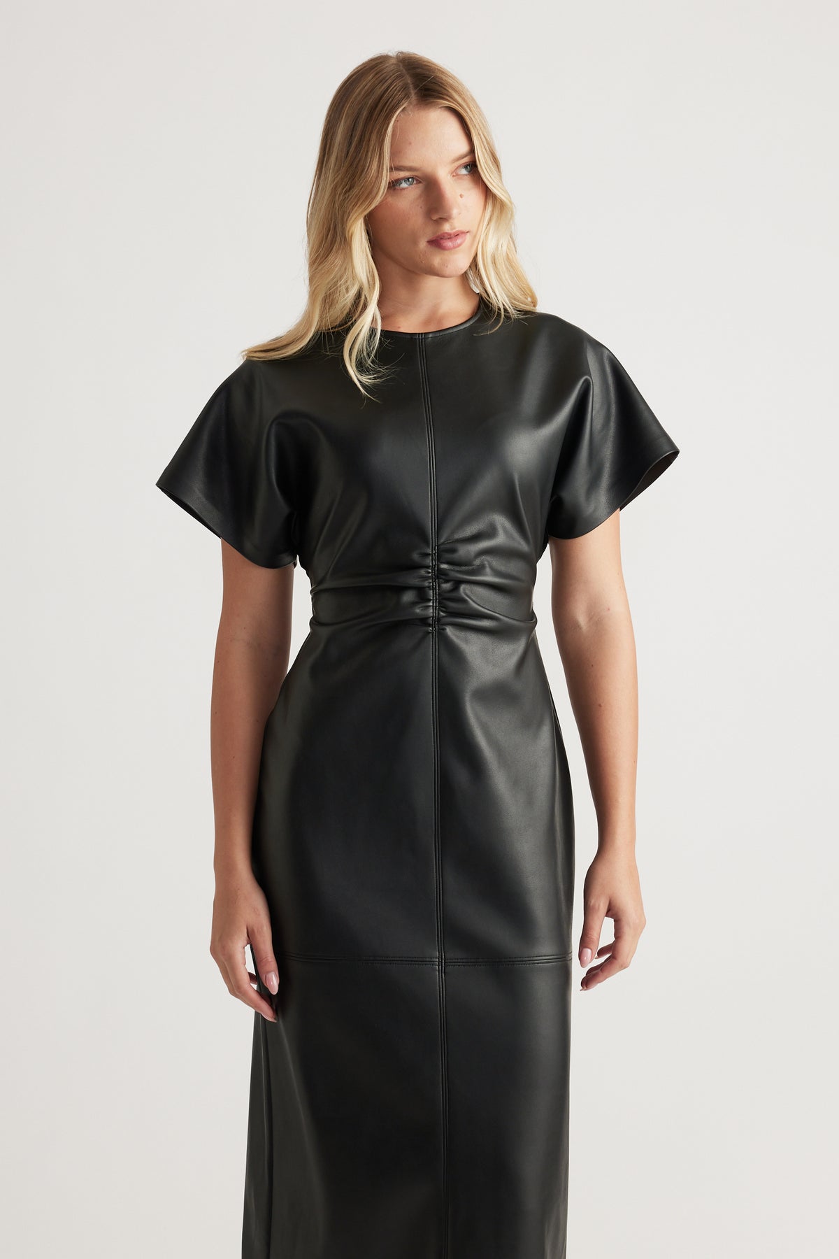 Woman wearing a black faux leather midi dress with short sleeves and gathered waist detailing against a neutral backdrop