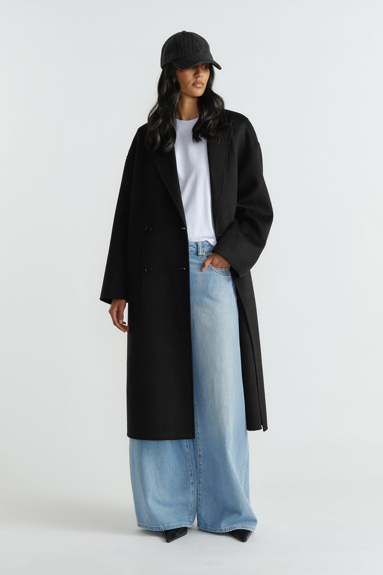 Woman wearing a long black tailored coat over a white tee, styled with wide-leg light wash denim jeans and a black cap