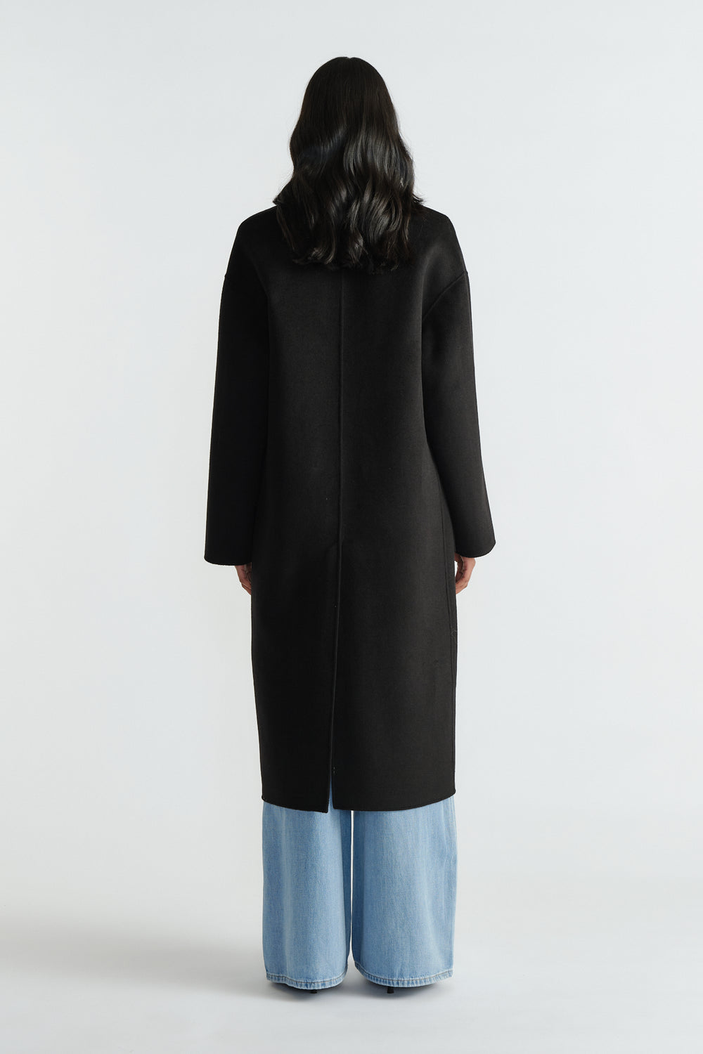 Back view of a woman wearing a sleek black long coat paired with wide-leg light blue denim jeans for a modern look