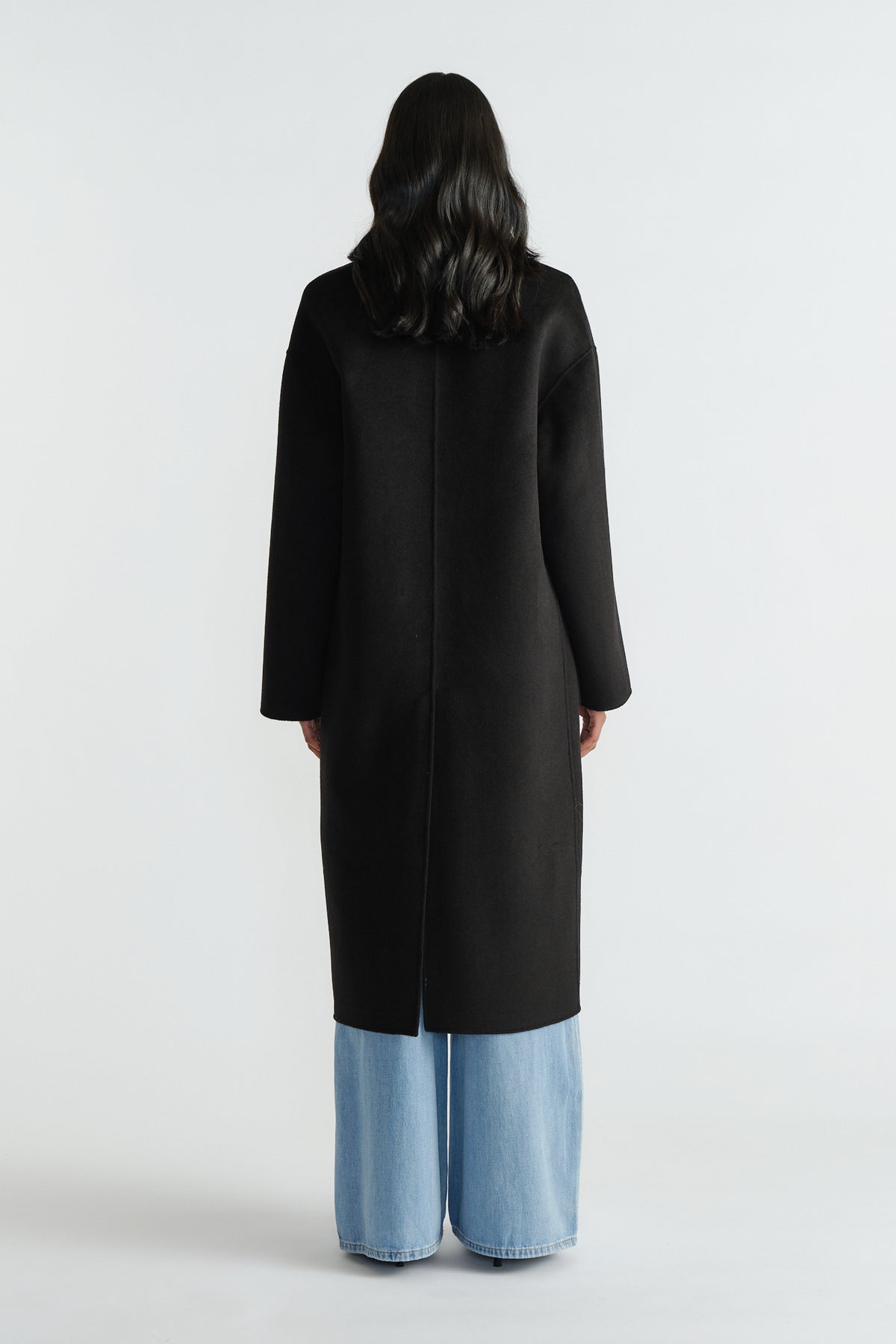Back view of a woman wearing a sleek black long coat paired with wide-leg light blue denim jeans for a modern look