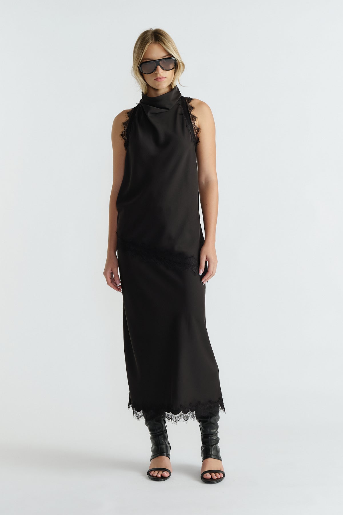 Woman wearing a sleek black slip dress with lace trim at shoulders and hem, styled with black boots and sunglasses