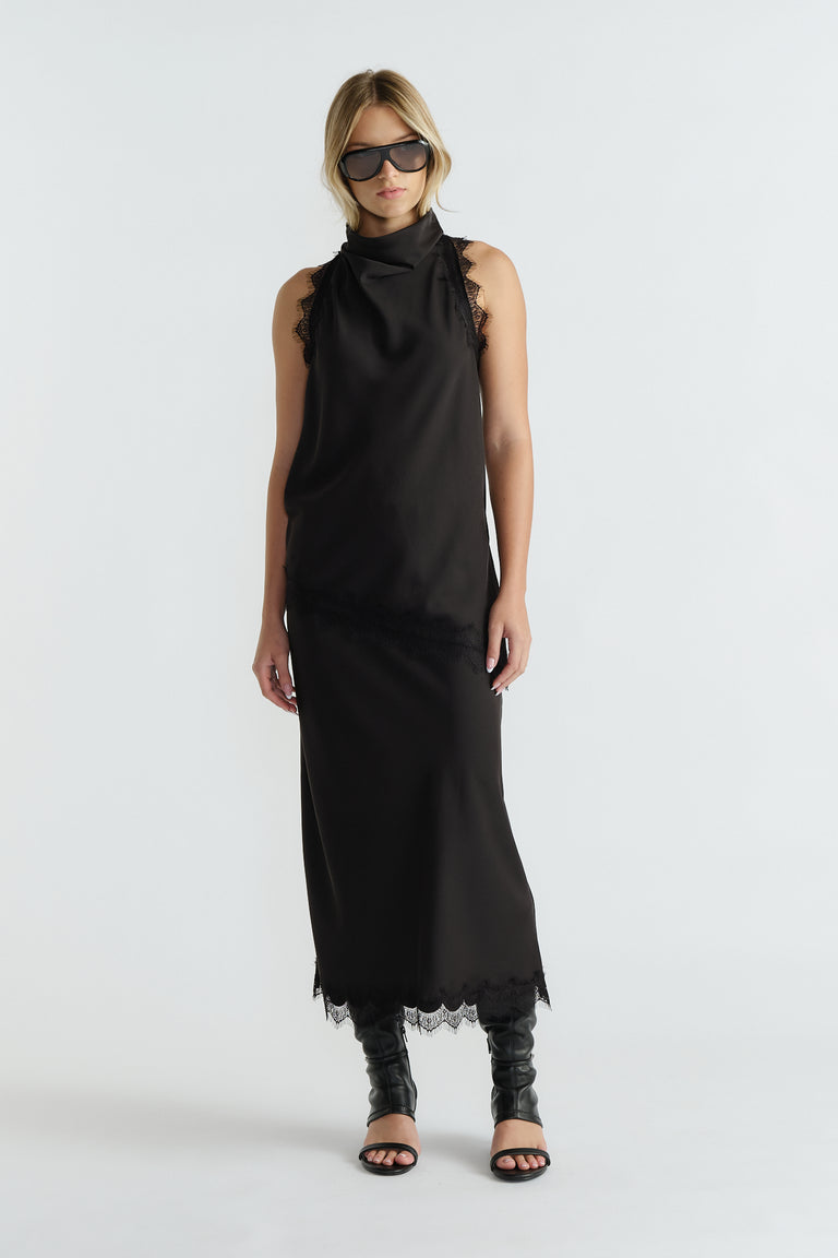 Woman wearing a sleek black slip dress with lace trim at shoulders and hem, styled with black boots and sunglasses