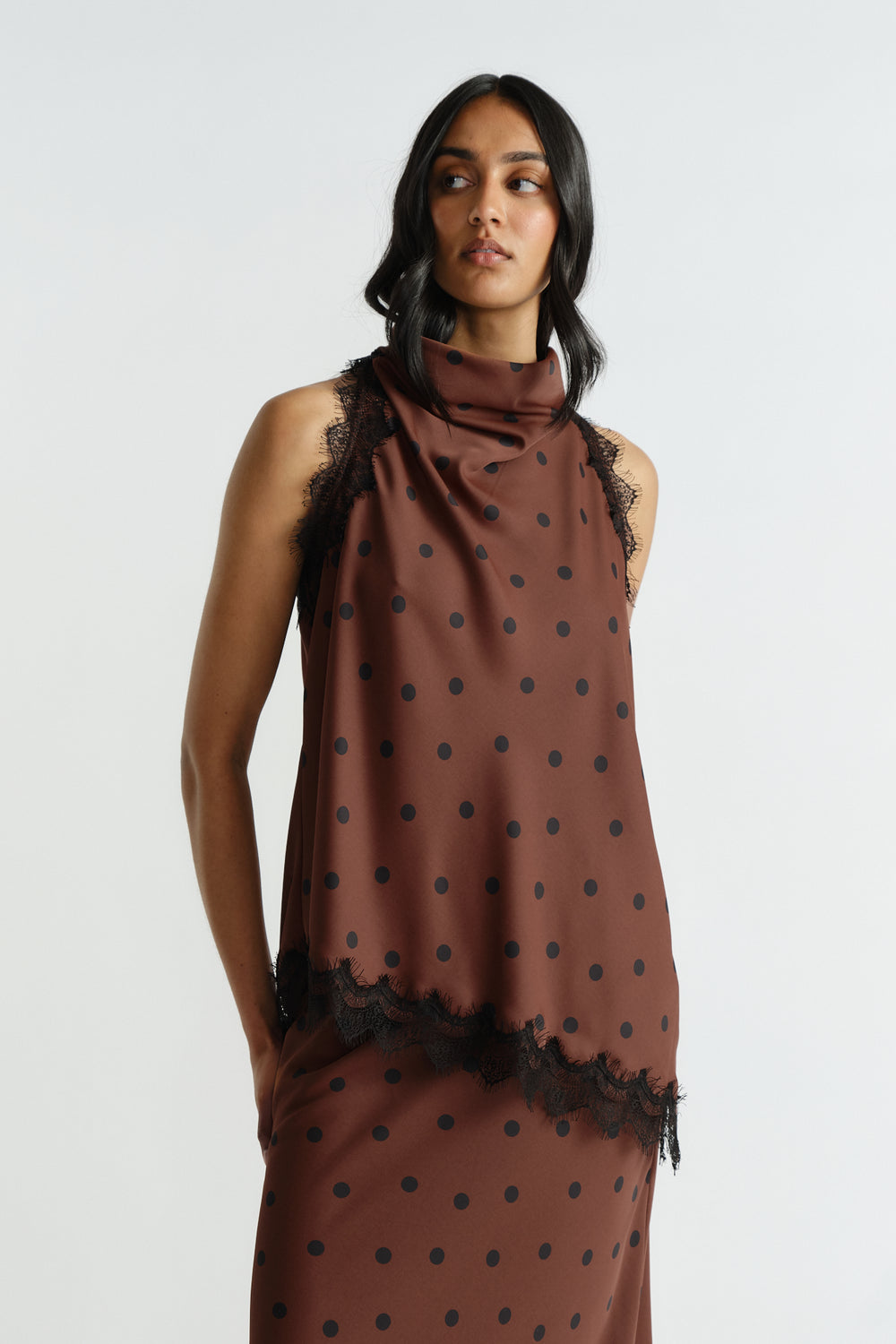 Woman wearing a sleeveless brown polka dot top with delicate black lace trim and a high draped neckline