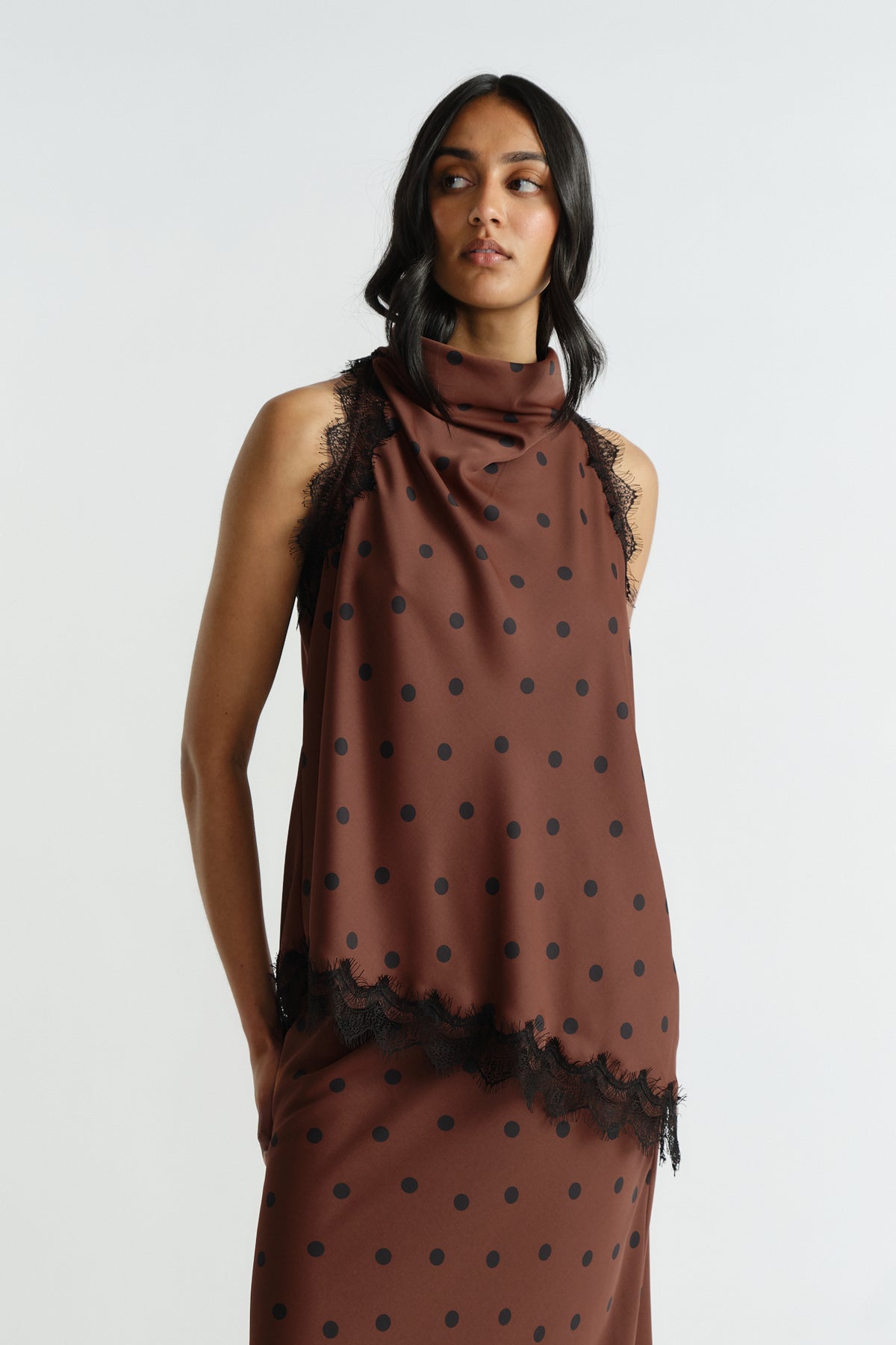 Woman wearing a sleeveless brown polka dot top with delicate black lace trim and a high draped neckline