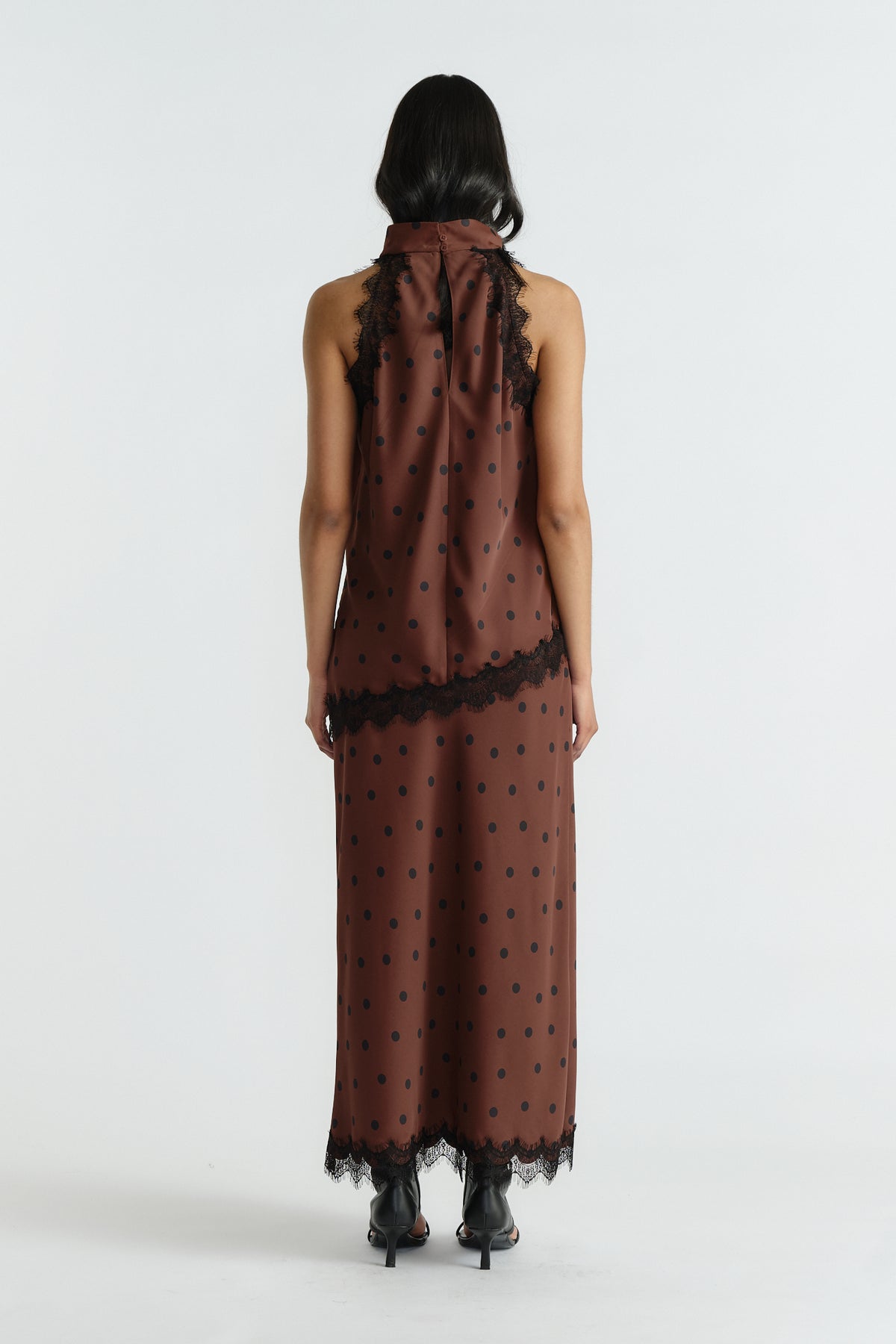 Back view of woman wearing a sleeveless brown polka dot halter top and matching skirt with black lace trim and heels