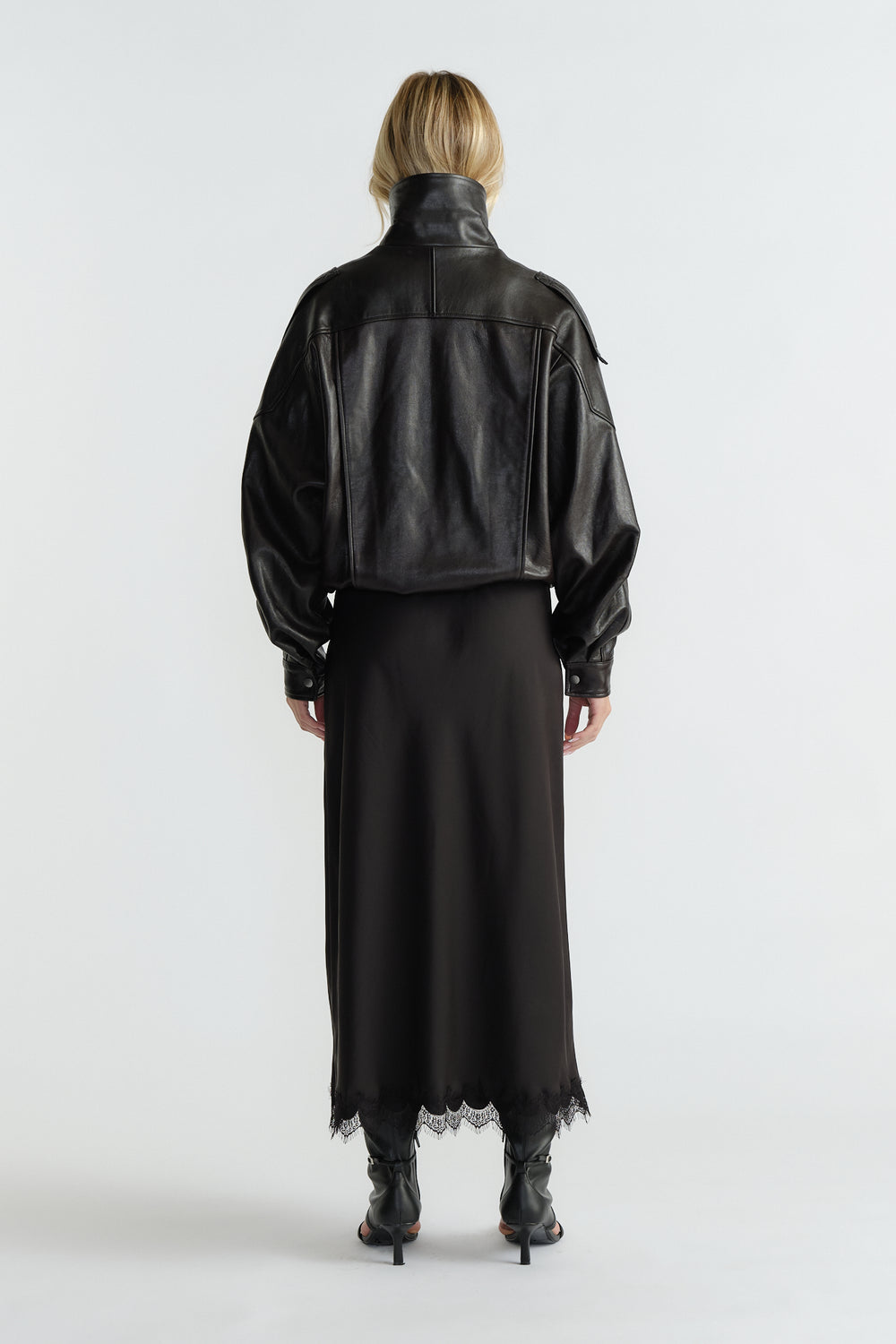 Back view of woman wearing a cropped black leather jacket paired with a flowing lace-trimmed maxi skirt and heeled boots