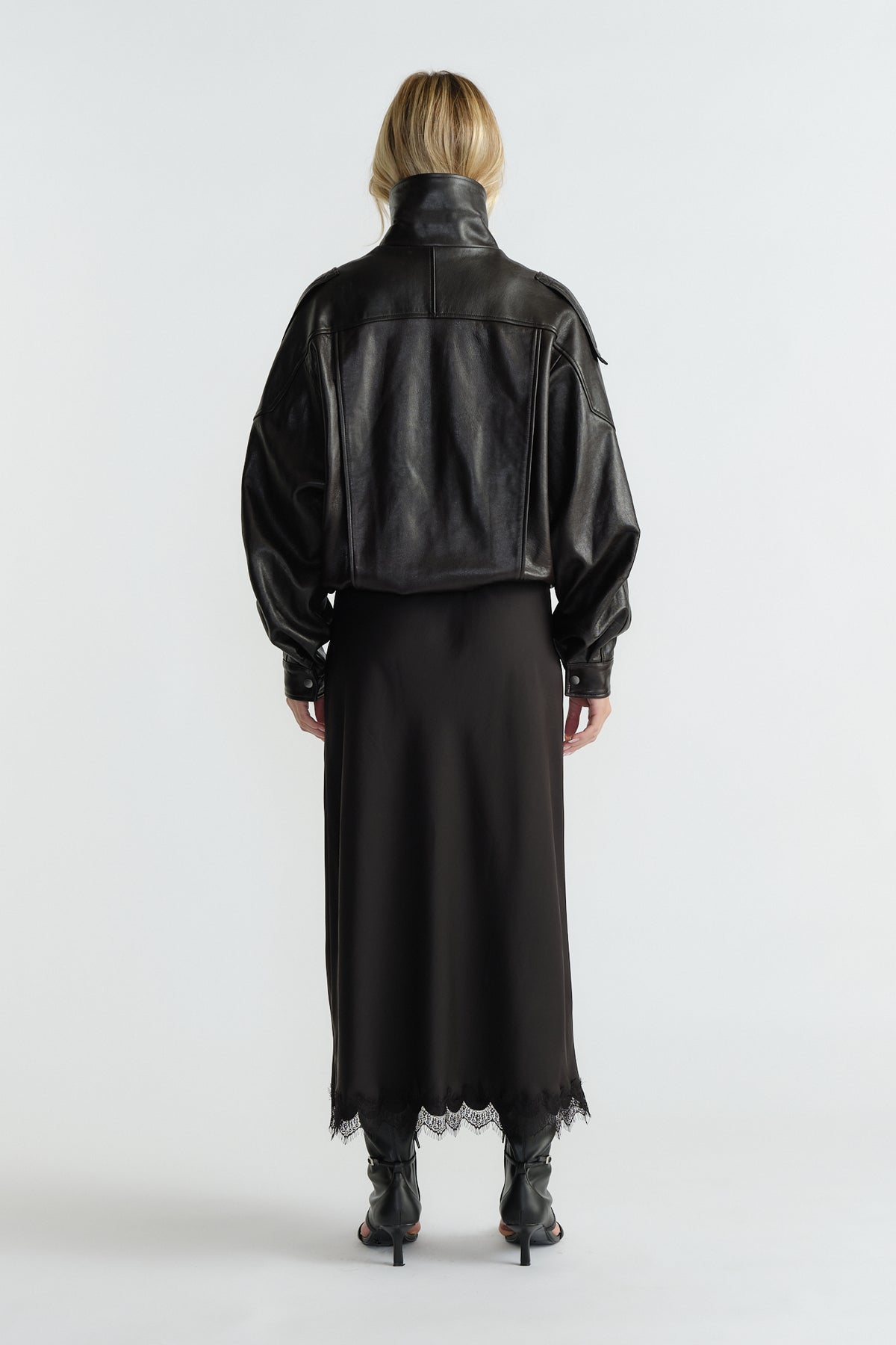 Back view of woman wearing a cropped black leather jacket paired with a flowing lace-trimmed maxi skirt and heeled boots