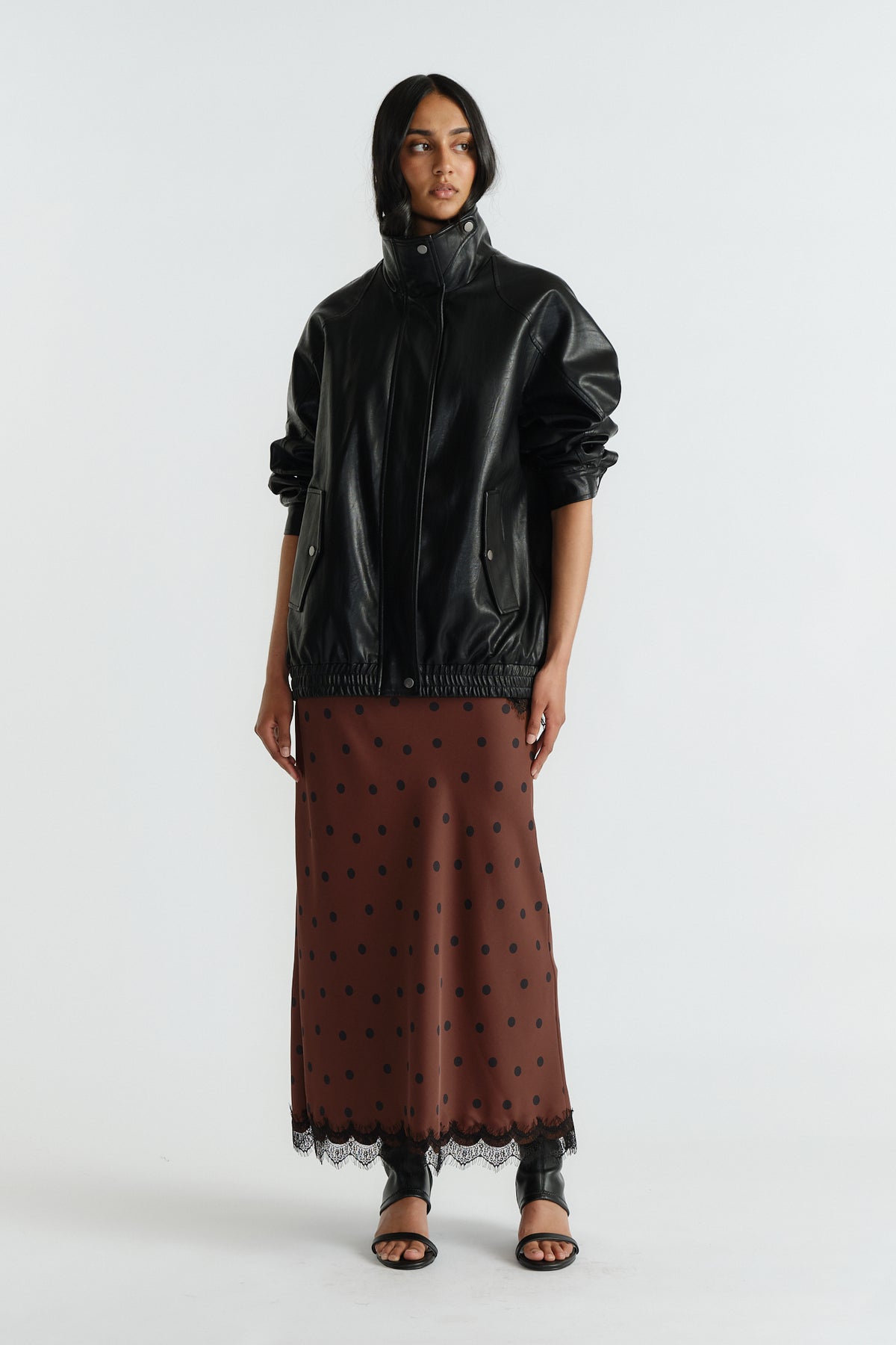 Woman dressed in a black faux leather jacket over a brown polka dot maxi skirt with lace trim and black sandals