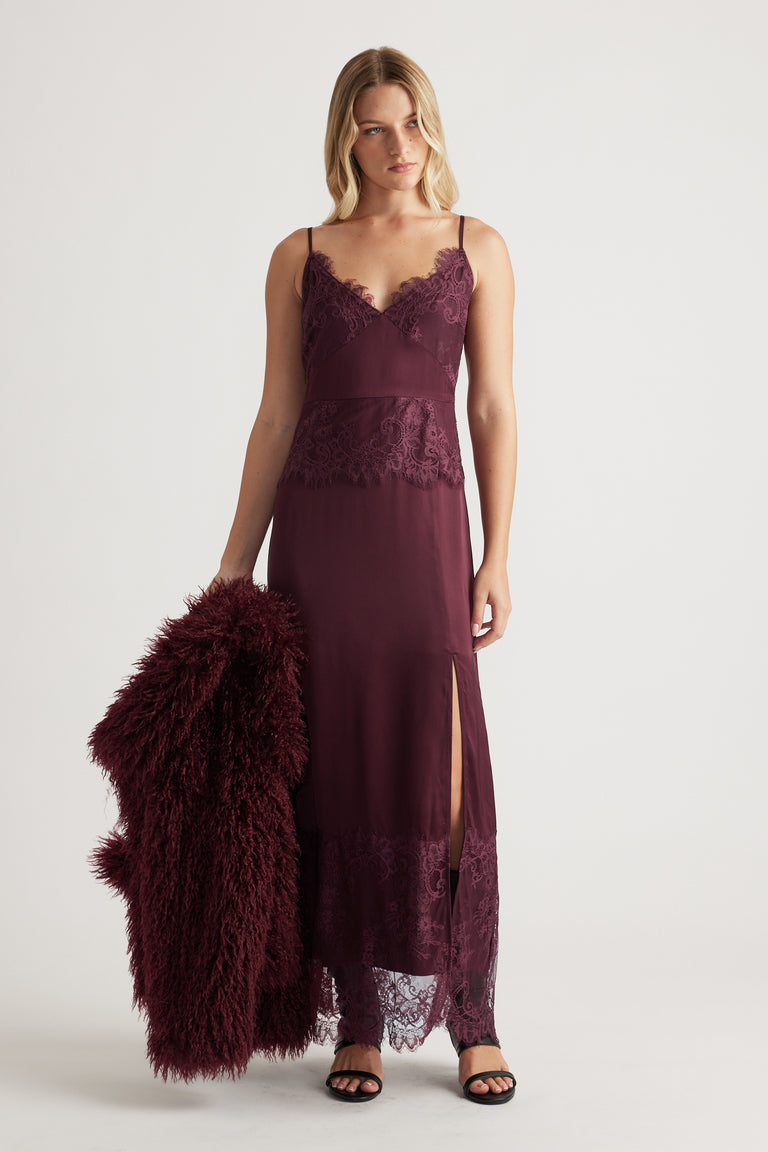 Woman wearing a plum lace maxi dress with thin straps and a high slit, holding a matching textured coat and black sandals