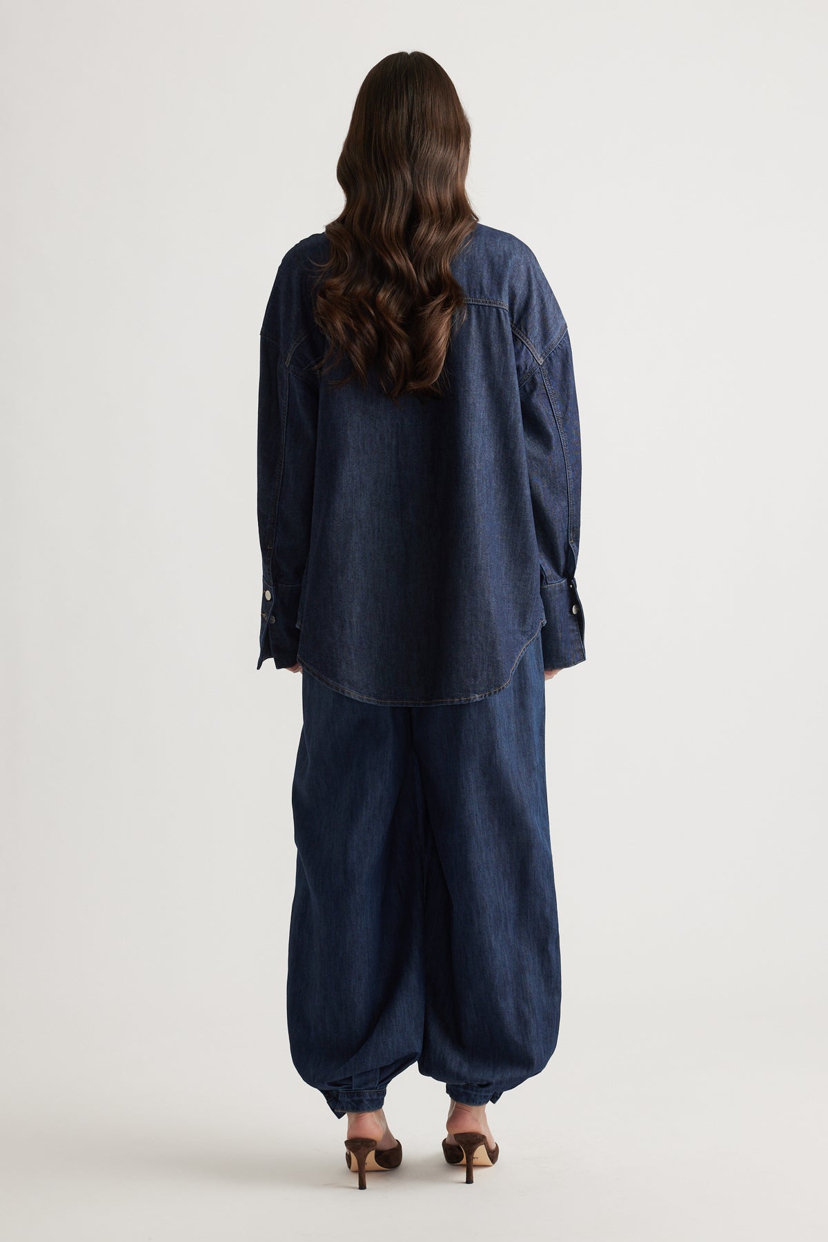 Woman seen from behind wearing a loose dark denim shirt paired with wide-leg denim pants and brown heeled sandals