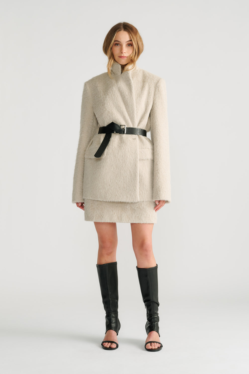Woman wearing a soft stone textured belted blazer layered over a matching mini skirt with black open-toe knee-high boots.