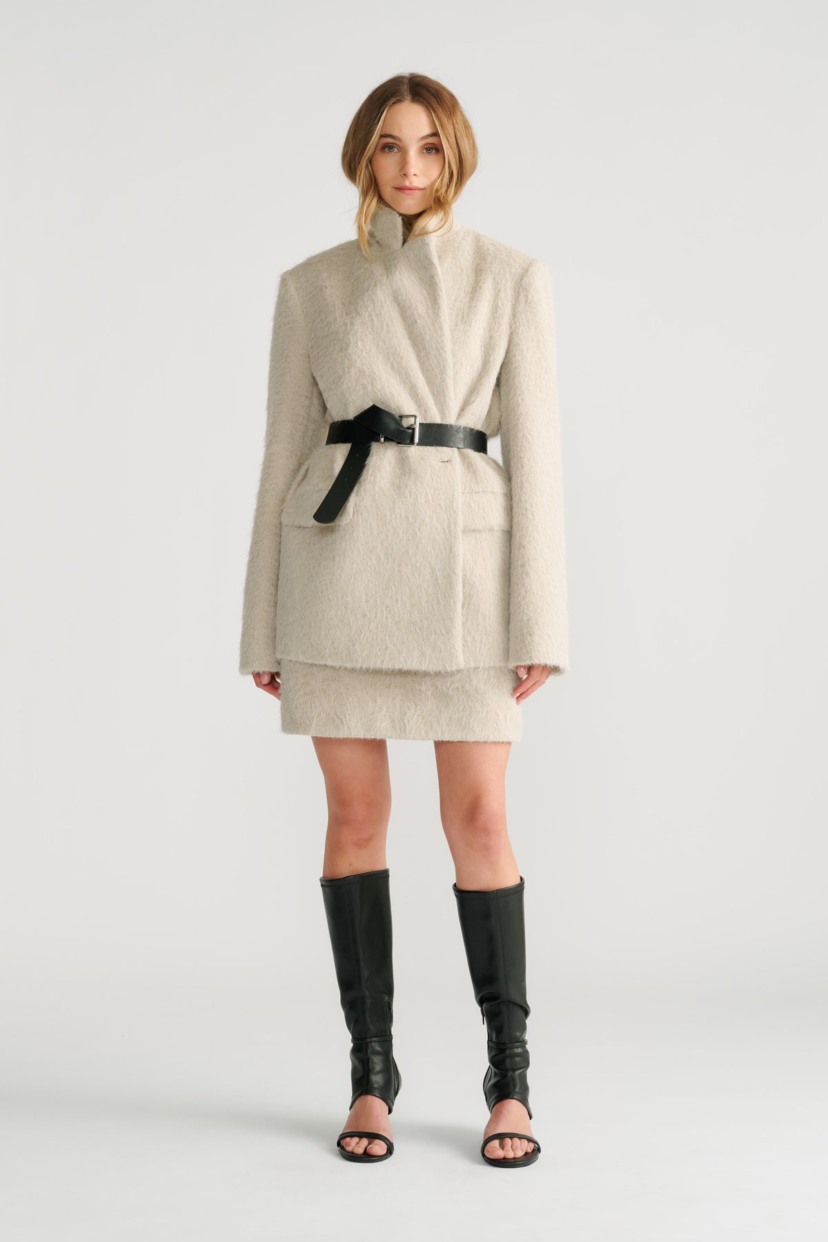 Woman wearing a soft stone textured belted blazer layered over a matching mini skirt with black open-toe knee-high boots.