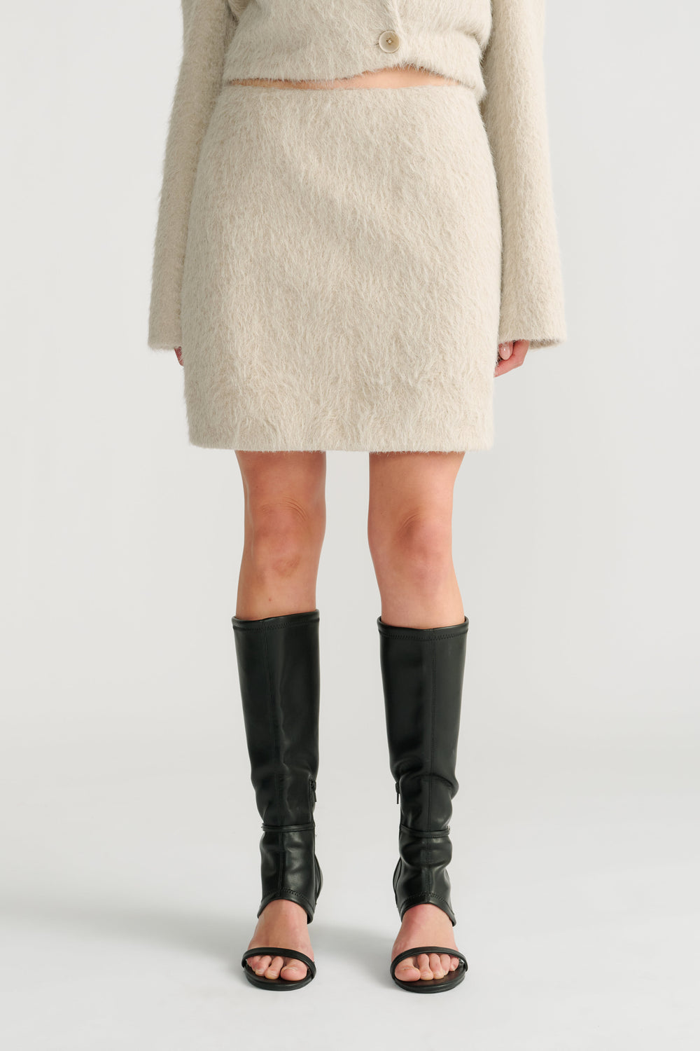 Woman wearing a soft stone-colored textured mini skirt paired with black open-toe knee-high boots and a matching top.