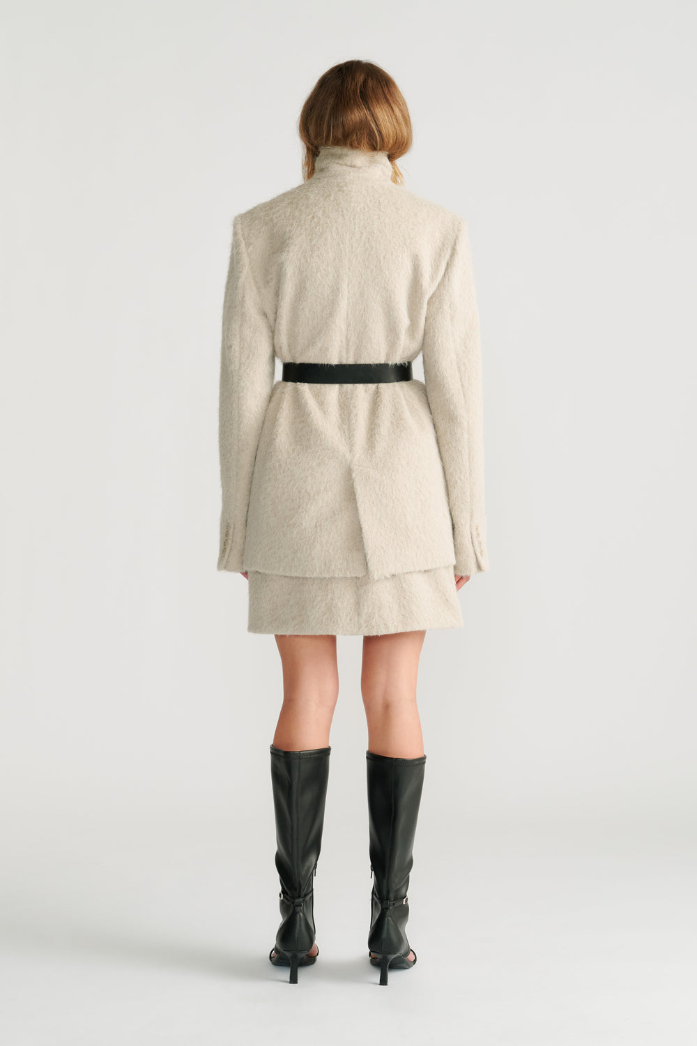 Woman wearing a soft stone textured belted jacket layered over a matching mini skirt with black high-heeled boots, back view.