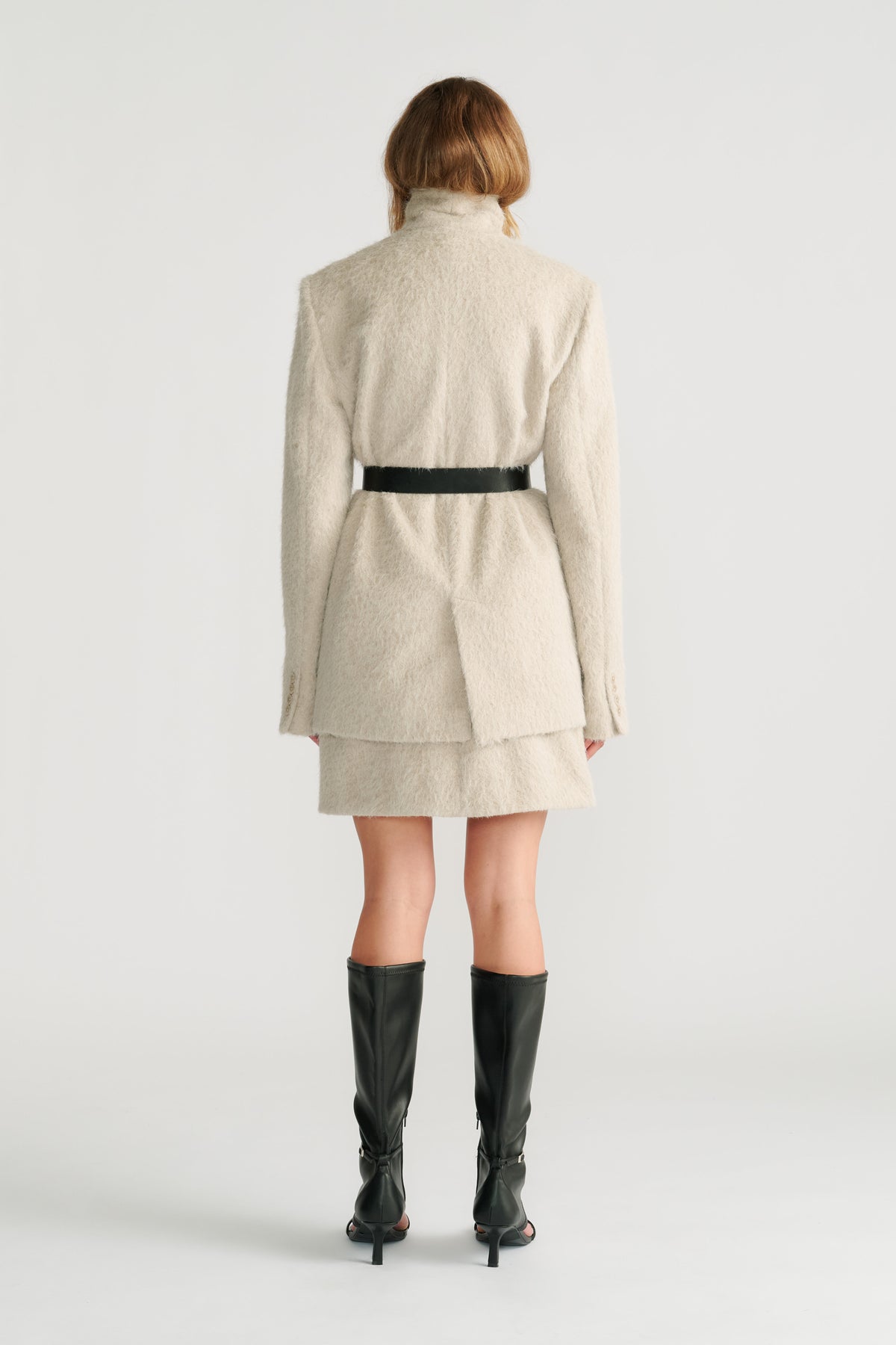 Woman wearing a soft stone textured belted jacket layered over a matching mini skirt with black high-heeled boots, back view.