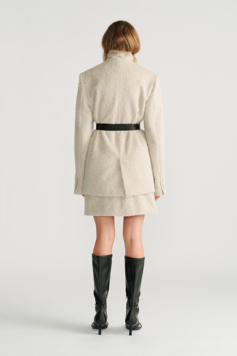 Woman wearing a soft stone textured belted jacket layered over a matching mini skirt with black high-heeled boots, back view.