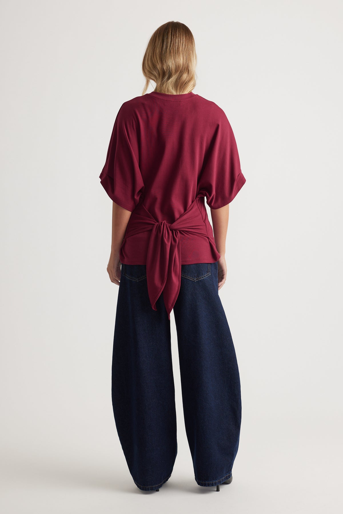 Back view of a wine-colored tee with a large tied bow detail, paired with wide-leg dark denim jeans for a relaxed style