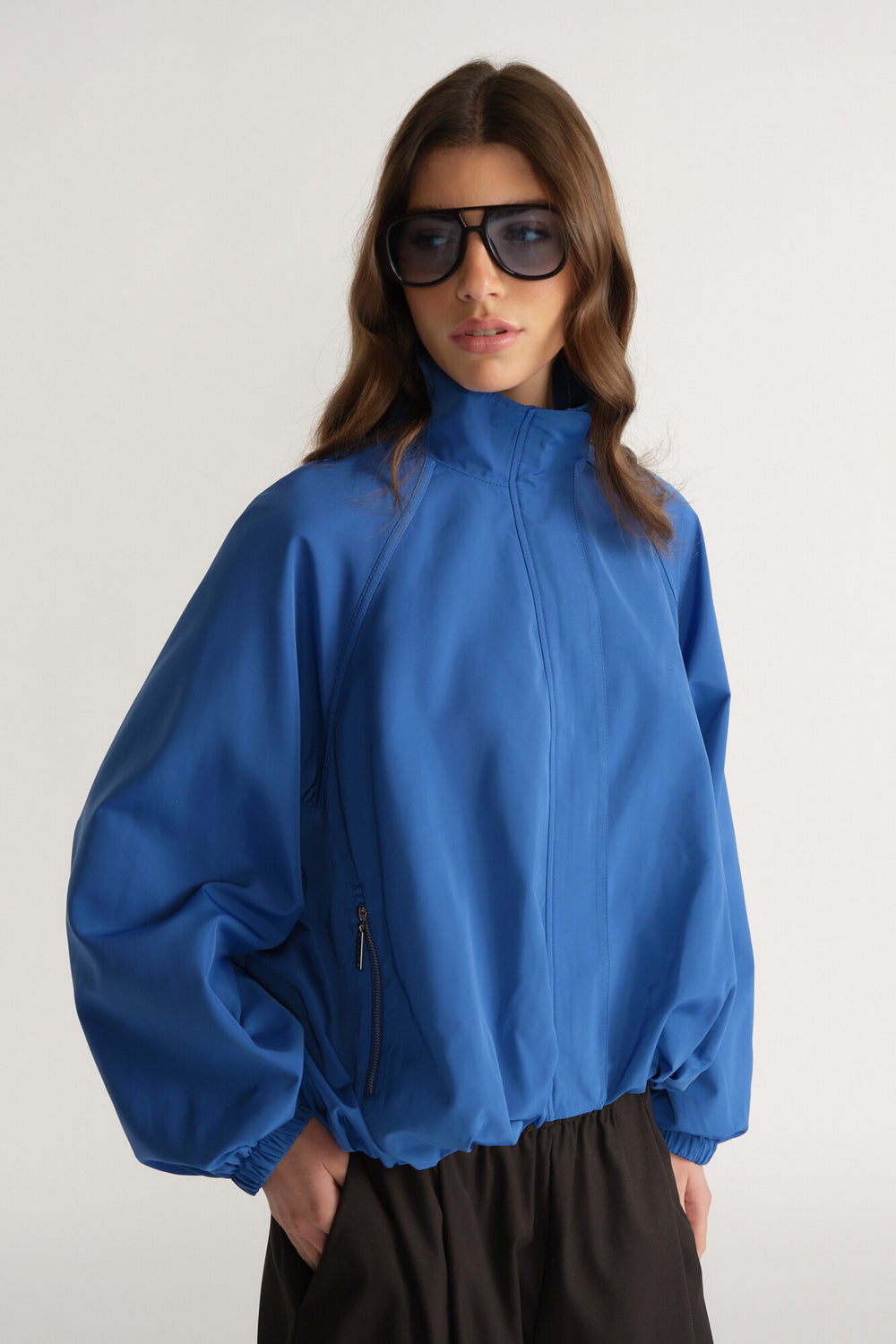 Woman wearing a vibrant blue oversized jacket with raglan sleeves and zippered pockets, styled with dark pants and sunglasses