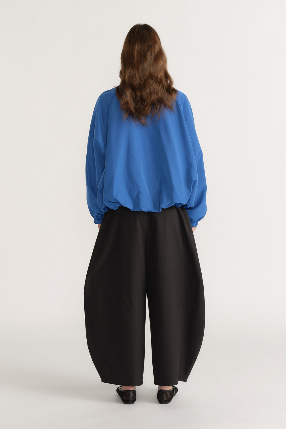 Woman seen from back wearing a loose blue long-sleeve top and wide-leg black pants with flat shoes