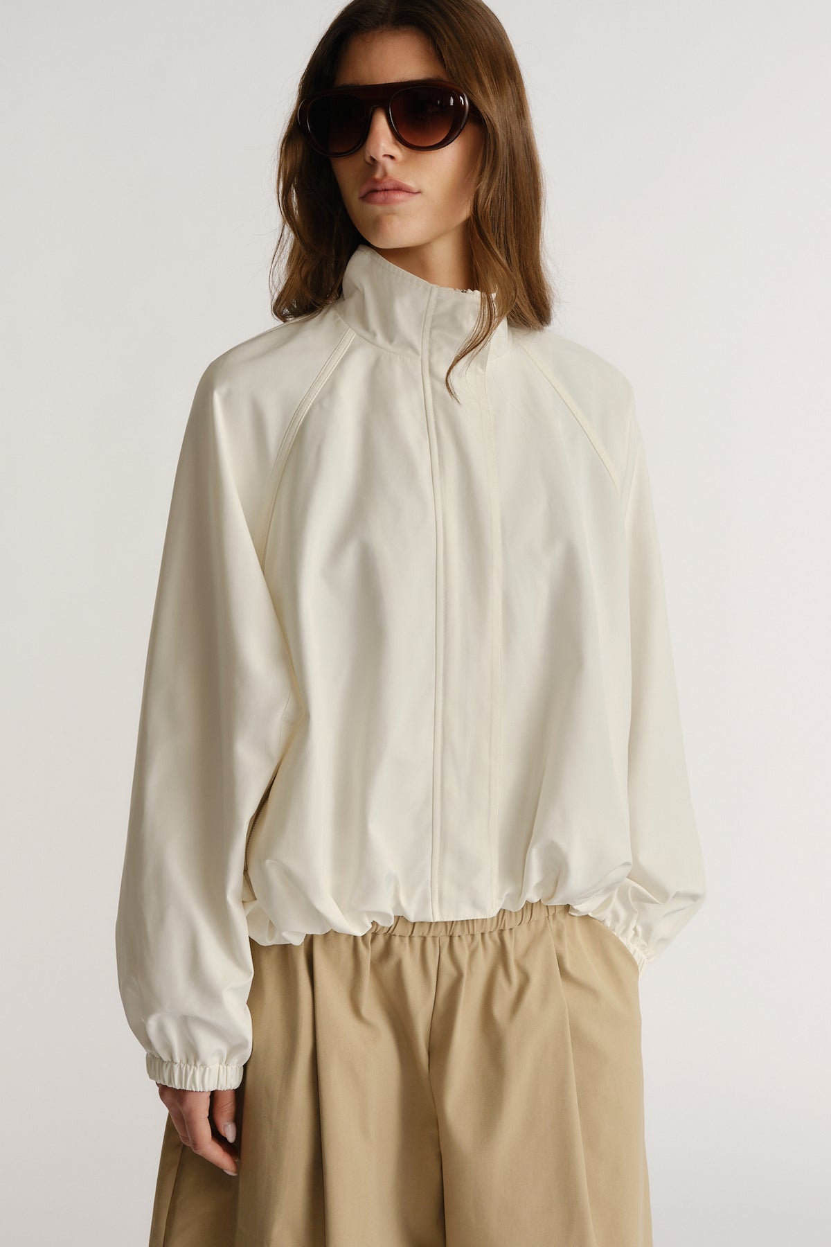 Woman wearing an ivory lightweight jacket with elastic cuffs and hem, styled with loose khaki pants and oversized sunglasses