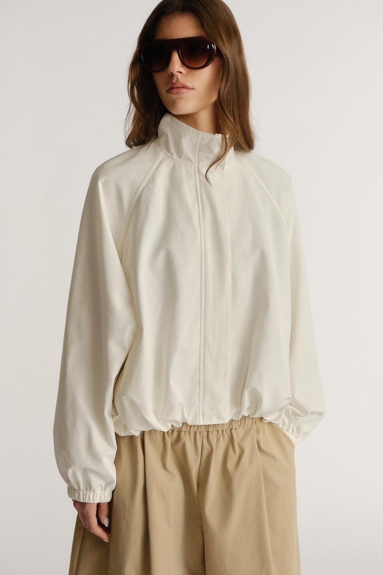 Woman wearing an ivory lightweight jacket with elastic cuffs and hem, styled with loose khaki pants and oversized sunglasses