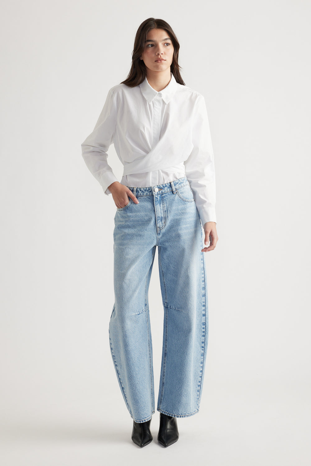 Woman wearing light blue wide-leg jeans with a white collared shirt and black pointed boots against a plain background.